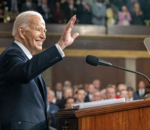 A smiling President Joe Biden at a podium waves to an assembled crowd in the US Capitol.